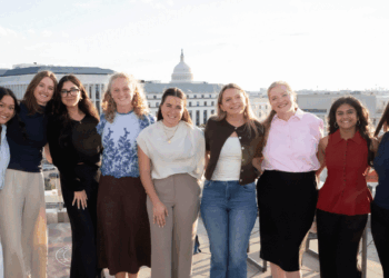 Nine women stand side by side, smiling on a rooftop with the U.S. Capitol building in the background on a sunny day.
