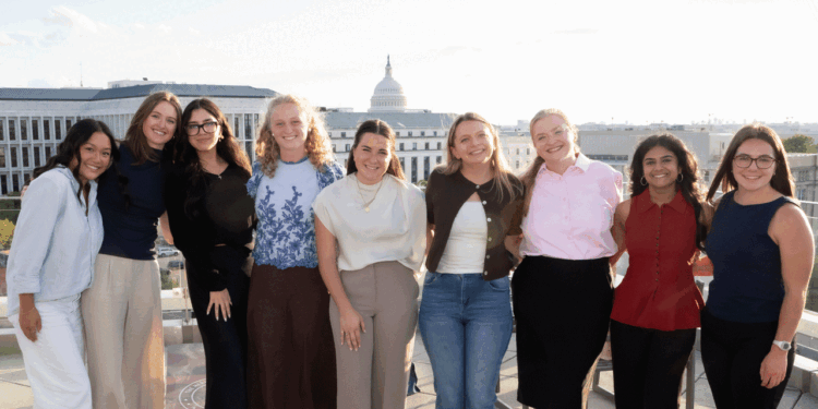 Nine women stand side by side, smiling on a rooftop with the U.S. Capitol building in the background on a sunny day.