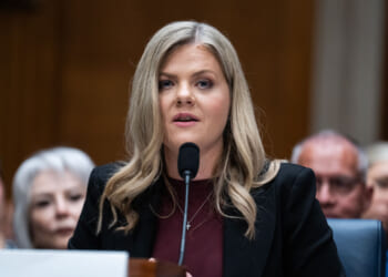 A woman with long blonde hair speaks into a microphone at a formal hearing, wearing a black blazer and maroon top. Several people sit blurred in the background.