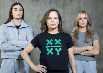 Three women stand confidently against a gray concrete wall. The center woman wears a black XX XY Athletics shirt; the other two wear light-gray athletic outfits with arms crossed. All look directly at the camera.