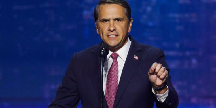 Todd Blanche in a dark suit and red tie speaks passionately at a microphone, gesturing with his right hand, against a blurred blue background.