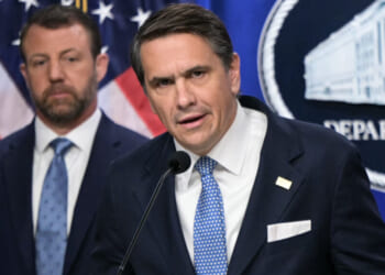 Todd Blanche and Markwayne Mullin in suits stand in front of U.S. flags and a sign that reads Department. The man in front speaks into a microphone, while the other looks on in the background.