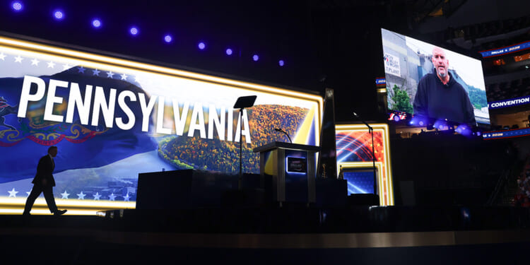 A stage with a large PENNSYLVANIA sign is lit up. A person stands in silhouette at the left. A screen shows another person speaking virtually, while empty seats are visible in the background.