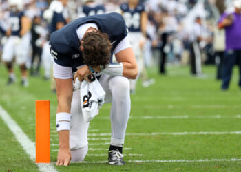 Football player in white pants and a dark jersey kneels on one knee near the end zone, head bowed in an expression of faith and hand touching the ground, with a towel in hand. Crowd and teammates are blurred in the background.