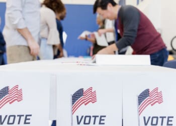 Ballots and American flags displayed on a table at a voting station, people are in the background.
