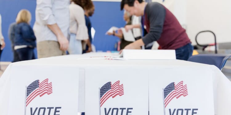 Ballots and American flags displayed on a table at a voting station, people are in the background.