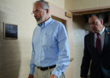 Two men walk through a hallway; one wears a light blue shirt and khaki pants, the other a dark suit and tie. The hallway has beige brick walls and a sign mounted on the left wall.