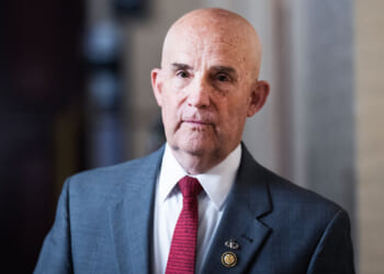 Bald man in a gray suit, red tie, and white shirt stands indoors. He has a serious expression and wears a pin on his lapel. The background is softly blurred.