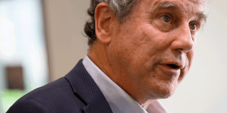 A man with gray hair wearing a dark blazer and light-colored shirt speaks, looking slightly to the side. The background is softly blurred, focusing attention on his face and expression.
