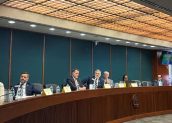 The Joint Election Equipment Specifications and Standards CommitteeA group of people meet at a long, curved conference table in a formal meeting room with microphones and nameplates in front of them. A TV screen shows another person in the background.