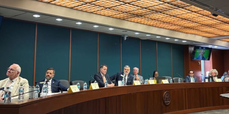 The Joint Election Equipment Specifications and Standards CommitteeA group of people meet at a long, curved conference table in a formal meeting room with microphones and nameplates in front of them. A TV screen shows another person in the background.