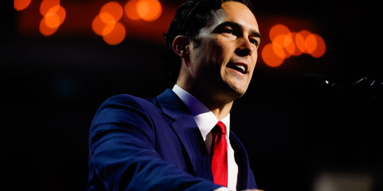 A man in a blue suit and red tie speaks at a podium against a dark background with orange lights blurred in the distance.