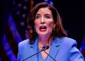 A woman in a blue blazer speaks at a podium with microphones, addressing the audience as she discusses the topic of pardon. She has shoulder-length brown hair and is shown against a dark background with blurred flags.