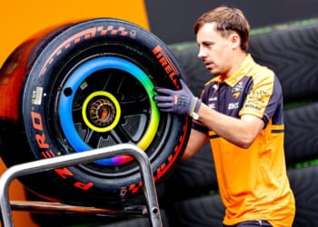 A mechanic in an orange and navy uniform carries a large Formula 1 Pirelli tire with colorful markings, likely for a racing event.