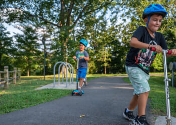 Two boys wearing helmets ride scooters on a paved path in a park surrounded by trees and grass on a sunny day.
