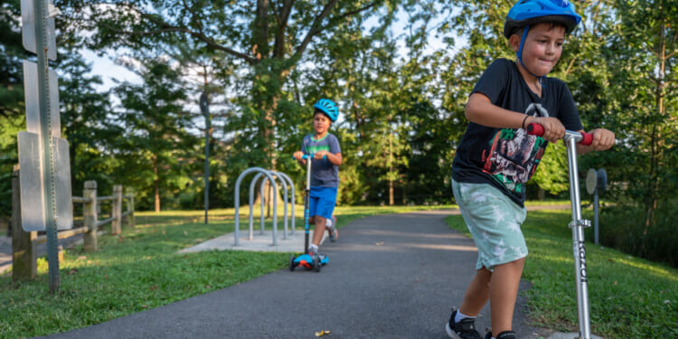 Two boys wearing helmets ride scooters on a paved path in a park surrounded by trees and grass on a sunny day.