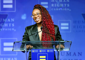 Human Rights Campaign President Kelley Robinson compared ICE to lynch mobs. Pictured: a person with long red braids, glasses, and a dark blazer speaks at a podium with a Human Rights Campaign logo against a blue background.