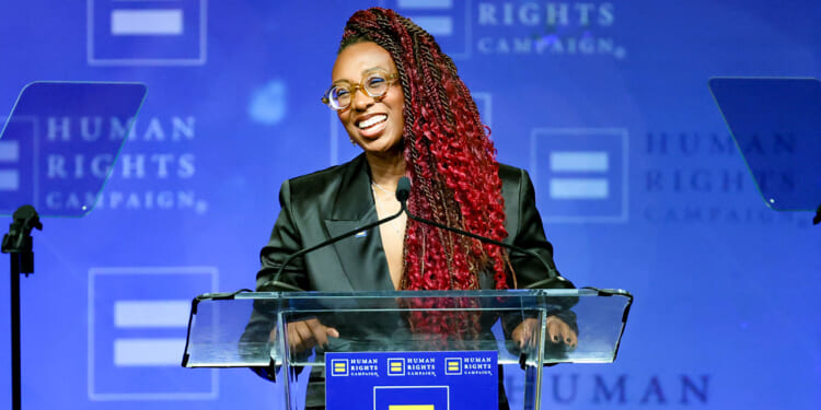 Human Rights Campaign President Kelley Robinson compared ICE to lynch mobs. Pictured: a person with long red braids, glasses, and a dark blazer speaks at a podium with a Human Rights Campaign logo against a blue background.