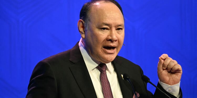 A man in a suit and tie speaks into microphones, gesturing with his right hand, against a blue background.