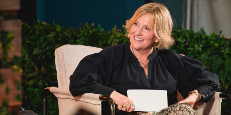 A woman with short blonde hair sits in a beige chair, smiling and holding a card. She wears a black blouse, floral skirt, and a headset microphone. Greenery and blurred background elements are visible behind her.