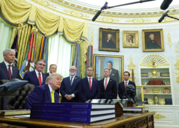 A group of men in suits, including Donald Trump seated at a desk, are gathered in the Oval Office with microphones overhead and shelves, flags, and presidential portraits in the background.