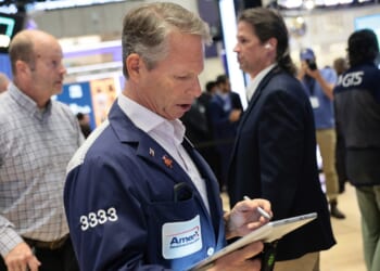 A stock trader in a blue jacket uses a tablet on a busy trading floor, surrounded by other people working and electronic screens displaying market data.