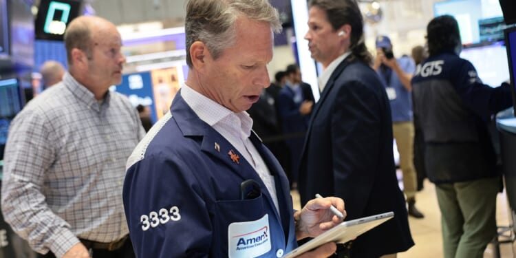 A stock trader in a blue jacket uses a tablet on a busy trading floor, surrounded by other people working and electronic screens displaying market data.