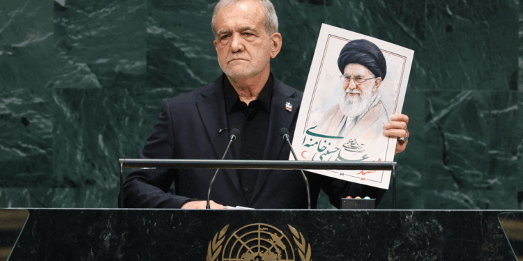 Iran's president stands at a United Nations podium holding a large portrait of a Ayatollah Khamanei wearing glasses and a turban, with Persian writing beneath the image, representing Iran. The UN emblem is visible on the podium.