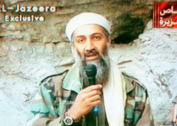 Osama Bin Laden in camouflage clothing and a white head covering speaks into a microphone against a rocky background, referencing the events of 9/11. Al-Jazeera Exclusive and Arabic text appear on the screen.