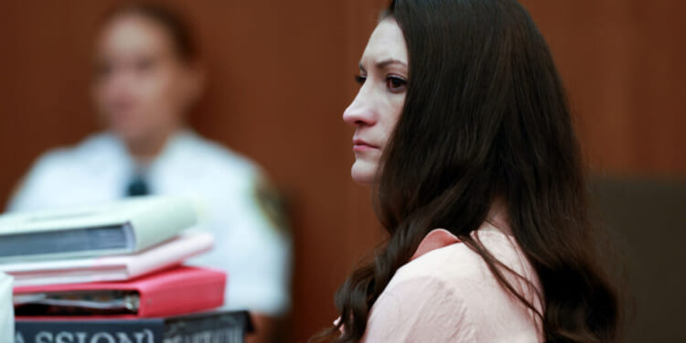 A woman with long brown hair sits in a courtroom, looking serious. Stacks of books and papers are in front of her, and a person in a white uniform is blurred in the background.