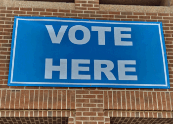 A blue sign with white letters reads VOTE HERE and is attached to a brick wall, indicating a polling location.