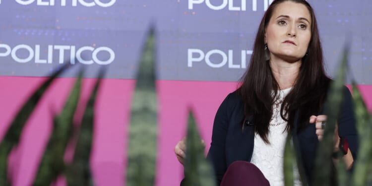 A woman with long brown hair sits on stage in front of a backdrop reading POLITICO, with green plants visible in the foreground.