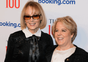 E. Jean Carroll and Roberta Kaplan smiling as they pose together at a red carpet event. One wears sunglasses and a black outfit with a bow, the other wears a white top with a black jacket. Logos and TIME 100 are visible on the backdrop behind them.
