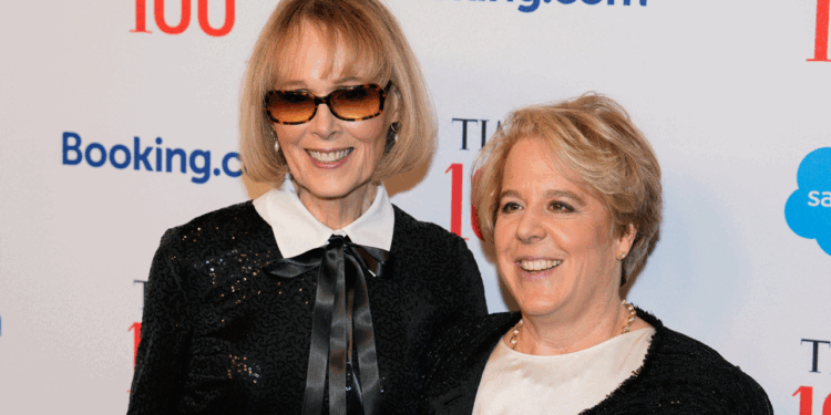 E. Jean Carroll and Roberta Kaplan smiling as they pose together at a red carpet event. One wears sunglasses and a black outfit with a bow, the other wears a white top with a black jacket. Logos and TIME 100 are visible on the backdrop behind them.