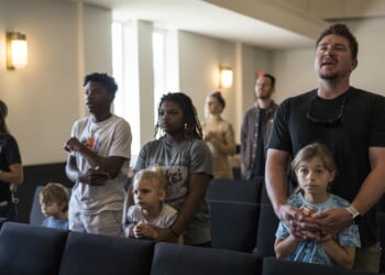 A group of adults and children stand together in a church pew, singing or listening intently during a service. The atmosphere is calm and attentive.