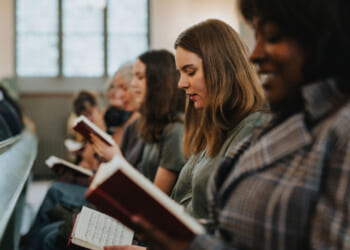 People sit in church pews reading hymnals or songbooks. The focus is on a young woman with brown hair, while others are also engaged in reading. The scene is well-lit, and a window is visible in the background.