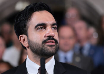 Zohran Mamdani in a suit with dark hair and a beard speaks at a podium, delivering an address. A blurred crowd is visible in the background.