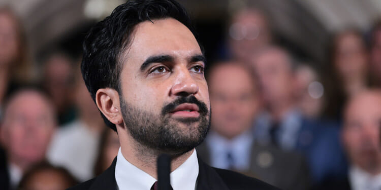 Zohran Mamdani in a suit with dark hair and a beard speaks at a podium, delivering an address. A blurred crowd is visible in the background.