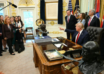 Donald Trump sits at the Oval Office desk, pointing while surrounded by officials, staff, and reporters. There are flags, documents, and busts visible in the ornate room. A photographer kneels, taking photos.