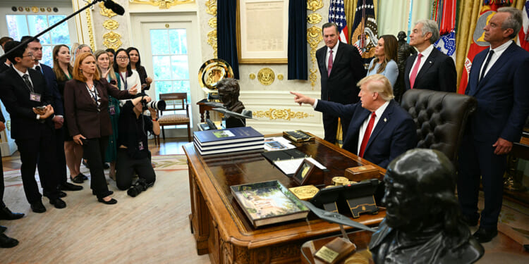 Donald Trump sits at the Oval Office desk, pointing while surrounded by officials, staff, and reporters. There are flags, documents, and busts visible in the ornate room. A photographer kneels, taking photos.