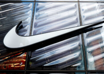 A large white Nike swoosh logo is displayed on a glass building facade, reflecting light and surrounding structures.