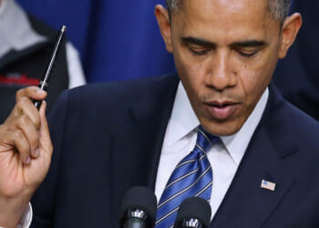 Barack Obama in a suit and striped blue tie, reminiscent of President Obama holds a pen up while speaking at a podium with microphones. An American flag pin is visible on the suit jacket.