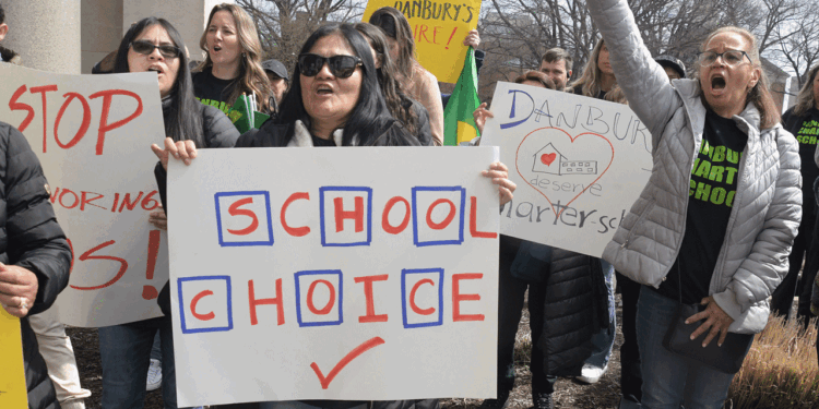 A group of people protest outdoors, holding signs with messages like SCHOOL CHOICE, STOP, and Danbury deserves a charter school. Some wear sunglasses and coats on a cloudy day.