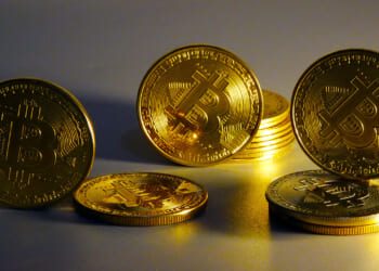 Several shiny gold coins with the Bitcoin symbol are arranged standing and lying on a flat surface, with a warm yellow light illuminating them.