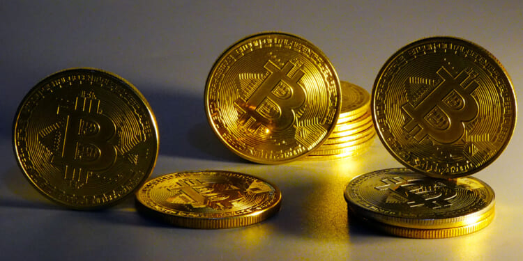 Several shiny gold coins with the Bitcoin symbol are arranged standing and lying on a flat surface, with a warm yellow light illuminating them.