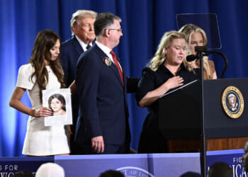 A woman speaks at a podium with the presidential seal, flanked by others. A man stands beside her and a young woman holds a photo of a child. President Donald Trump and woman are in the background.