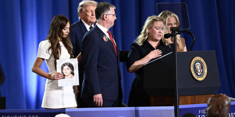 A woman speaks at a podium with the presidential seal, flanked by others. A man stands beside her and a young woman holds a photo of a child. President Donald Trump and woman are in the background.