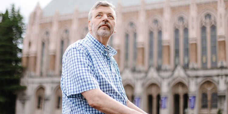 University of Washington professor Stuart Reges, who got disciplined over a land acknowledgment. Pictured: Reges, an older man in a blue plaid shirt sits outdoors, looking upwards thoughtfully. A large, historic building with arched windows and detailed stonework is in the blurred background.