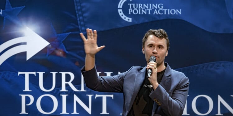 A man in a blazer speaks into a microphone, raising one hand, in front of a blue Turning Point Action backdrop with white text and arrow graphics, reminiscent of events frequently hosted by Charlie Kirk.