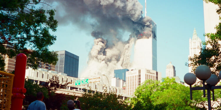 The South Tower is collapsing and smoke billows from the North Tower of the World Trade Center towers in New York City during daylight on Sept. 11, with people observing from a distance and surrounding buildings visible.